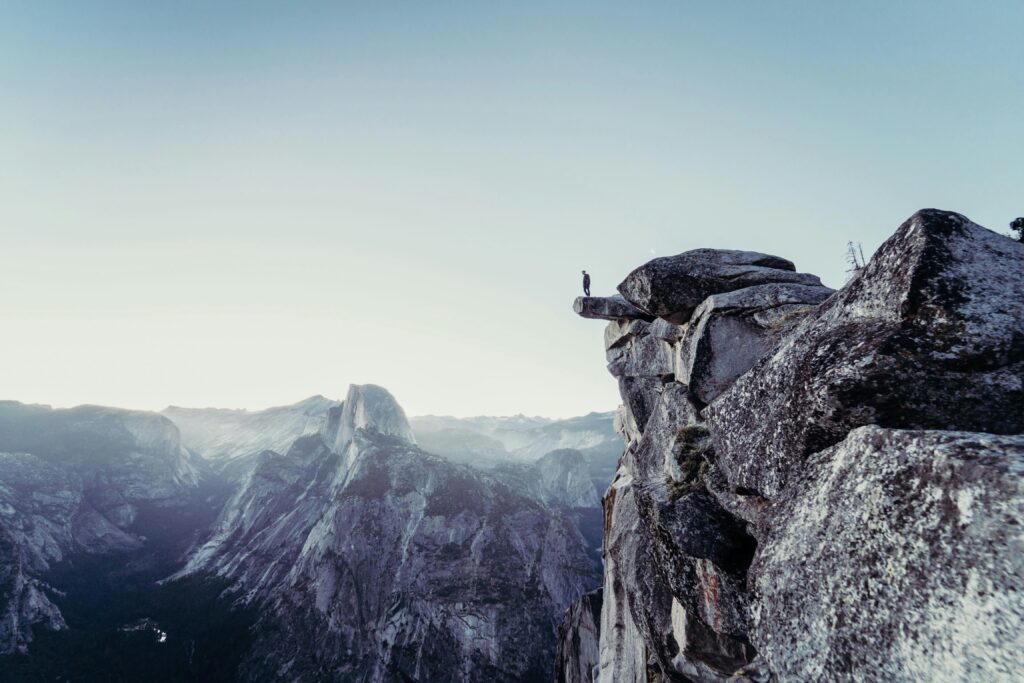 A man standing on the edge of a cliff in the mountains, overlooking a river flowing between peaks. The scene features hues of grey and blue, capturing the serene and dramatic landscape