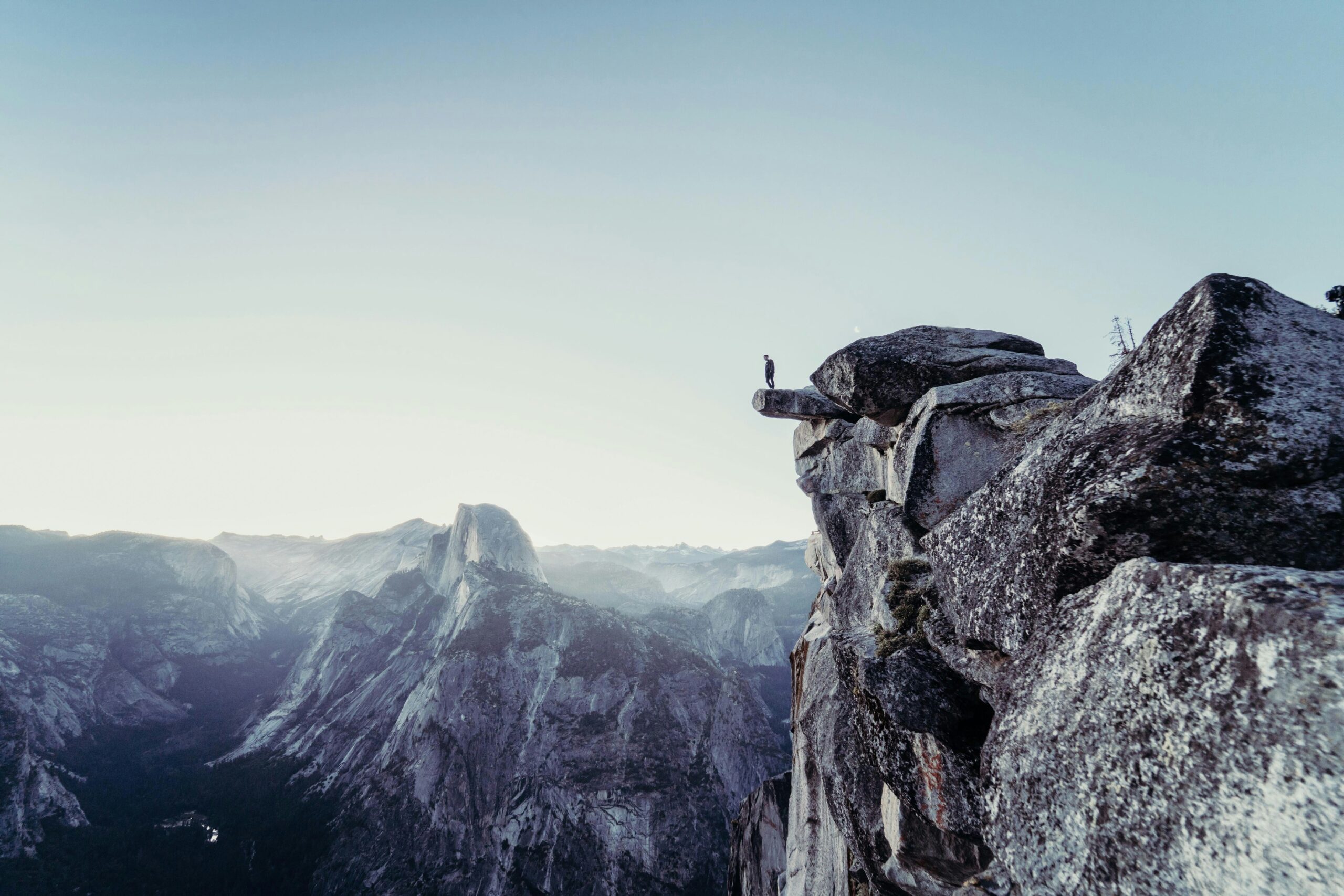A man standing on the edge of a cliff in the mountains, overlooking a river flowing between peaks. The scene features hues of grey and blue, capturing the serene and dramatic landscape