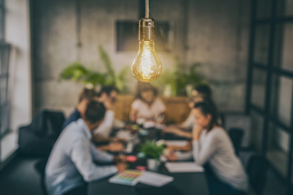 Unfocused image of employees seated at a conference table working on a project and filling in spreadsheets, with the camera focused on a light bulb emitting a warm glow.
