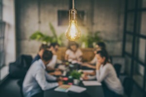 Unfocused image of employees seated at a conference table working on a project and filling in spreadsheets, with the camera focused on a light bulb emitting a warm glow.