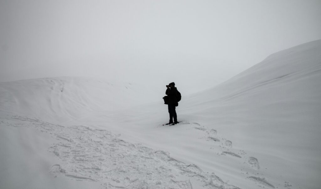 A man hiking in the tundra, capturing a photo of snowy hills and surrounding mountains. The landscape features stark white and black contrast, emphasizing the dramatic scenery.
