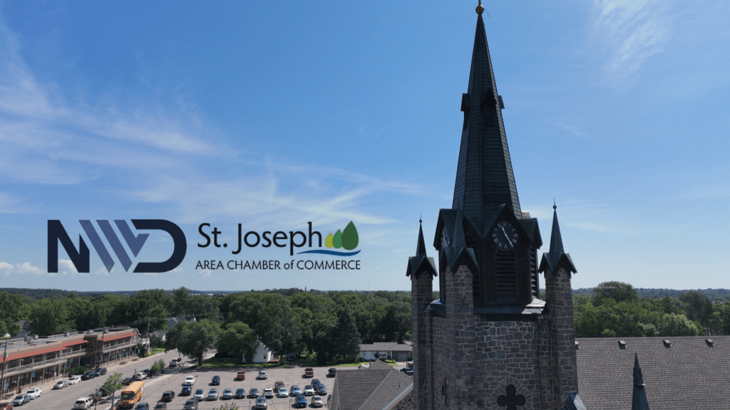 A Catholic church in St. Joseph, MN, featuring a tall steeple and historic architecture, with part of the main shopping area visible. The sky is light blue with wispy clouds.