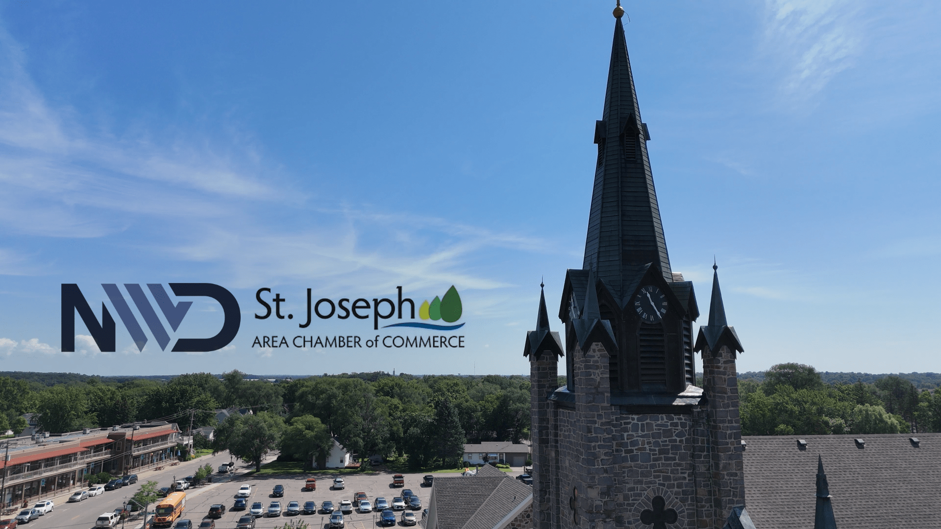 A Catholic church in St. Joseph, MN, featuring a tall steeple and historic architecture, with part of the main shopping area visible. The sky is light blue with wispy clouds.